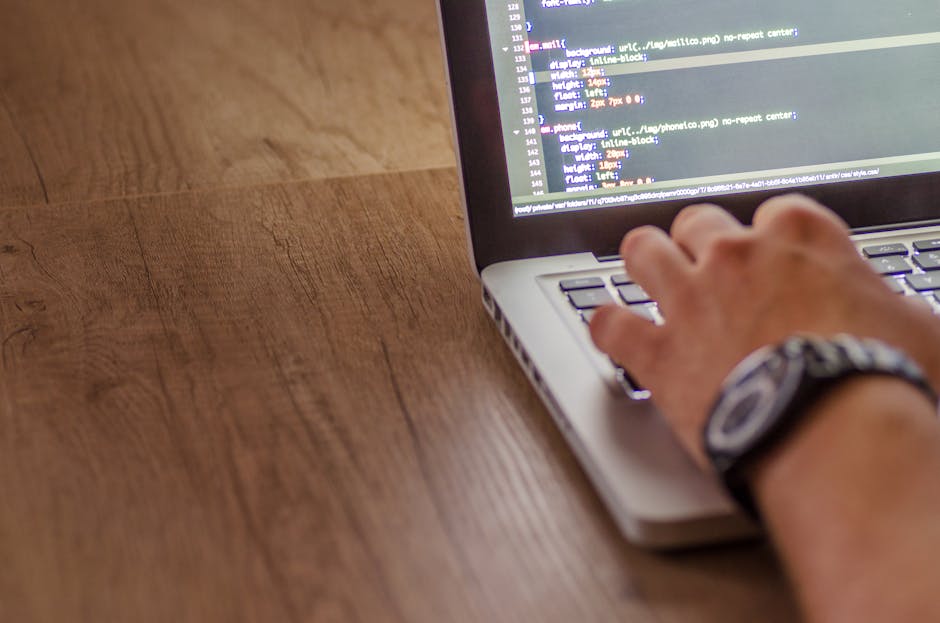 This image shows a person's hand wearing a wristwatch, typing on a laptop keyboard placed on a wooden desk. The screen displays lines of code or script, indicating programming or web development work.